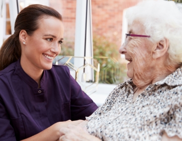 a woman kneels down and smiles beside a seated senior woman